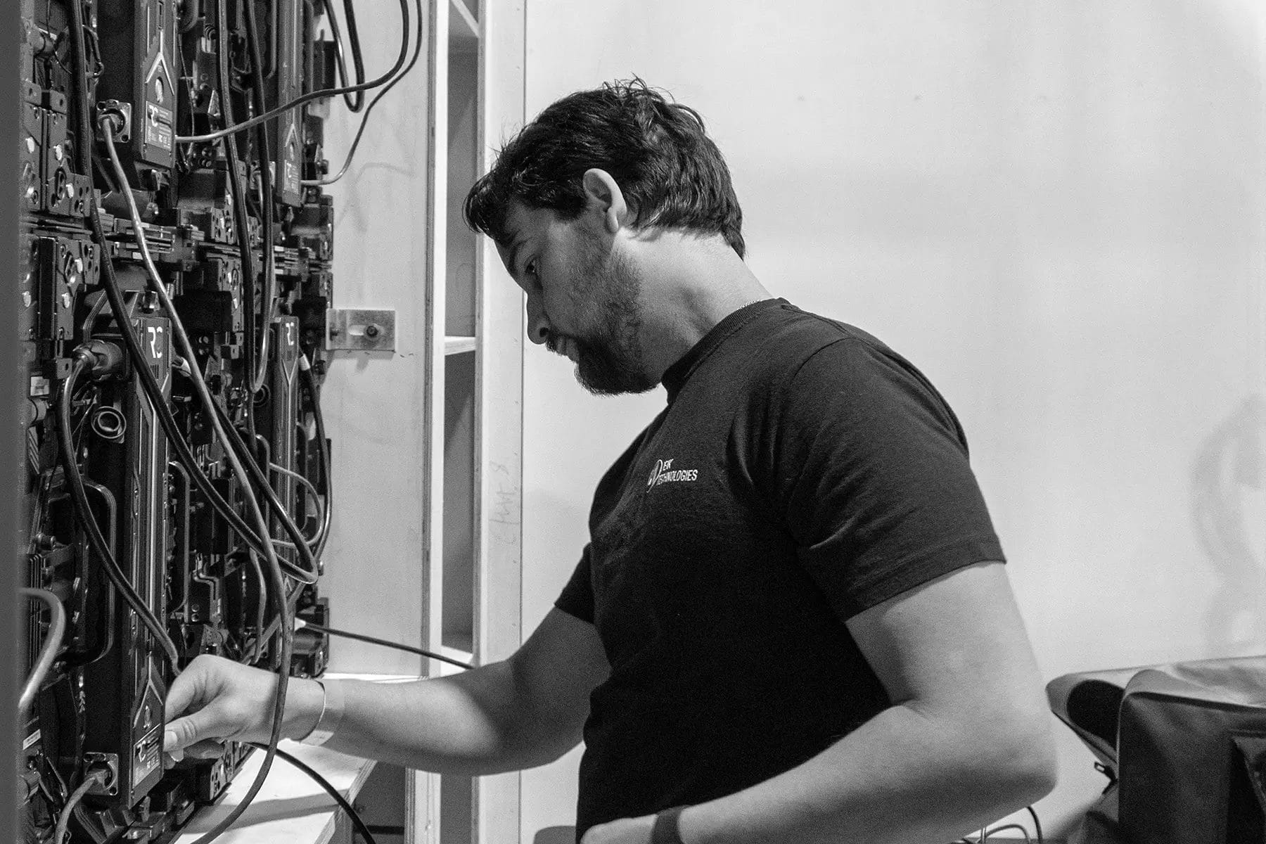 A man in a dark t-shirt works with cables and equipment in a server room, adjusting connections on a rack-mounted system. The image is in black and white.