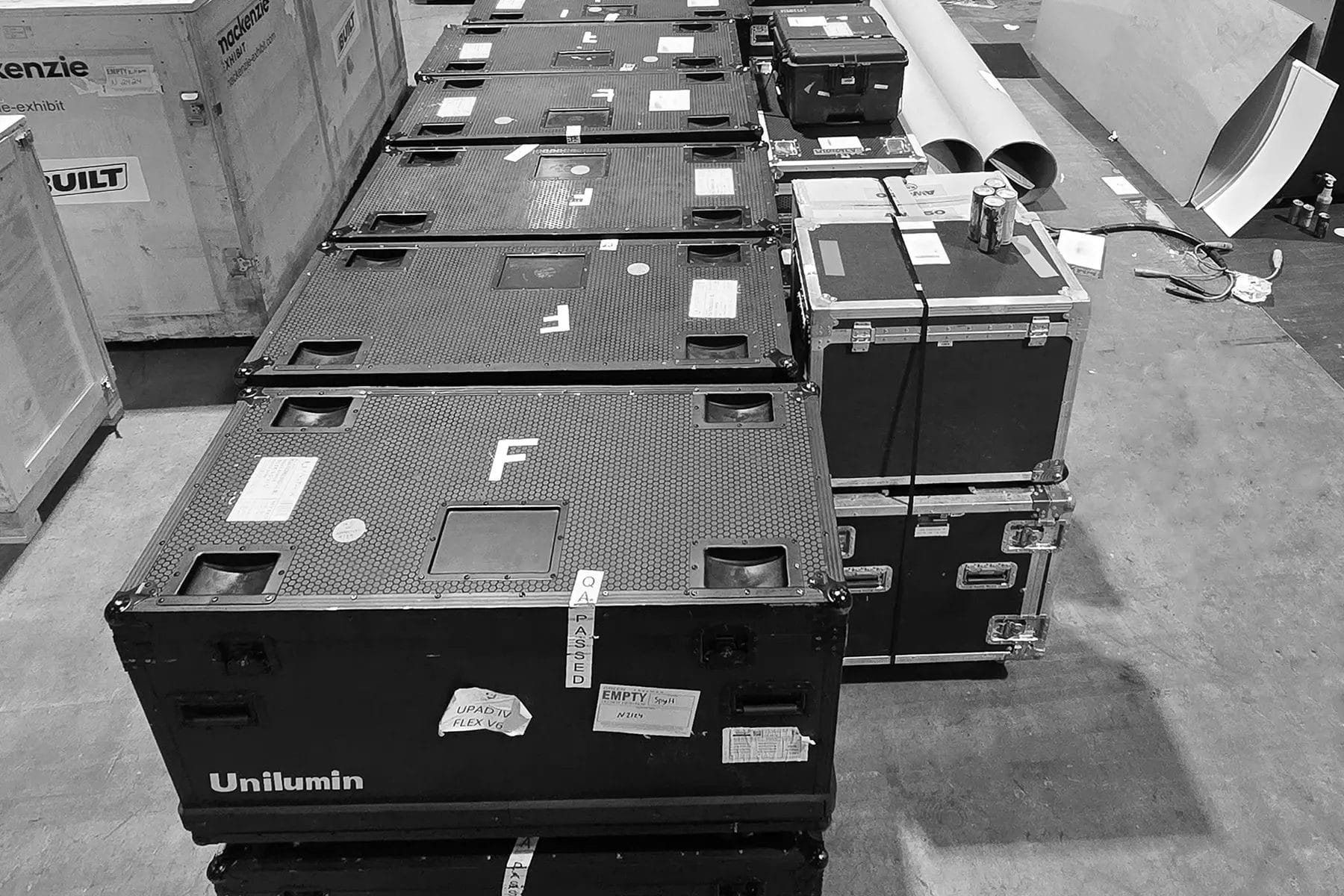 Several large black storage cases, some labeled "Unilumin," are lined up on a warehouse floor, surrounded by boxes, cardboard tubes, and other equipment. The scene appears organized and industrial.
