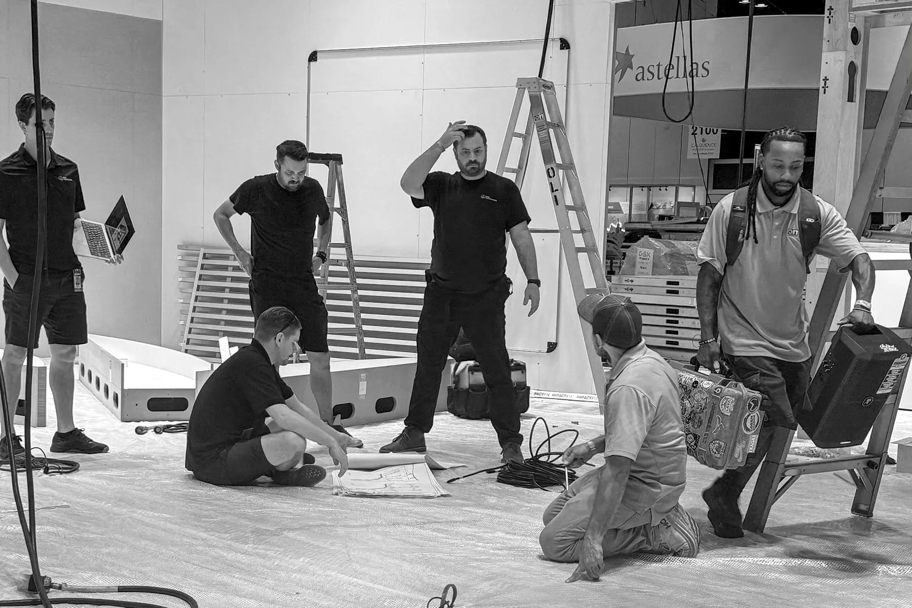 Six men are setting up an exhibit booth; some stand, some kneel or sit. One man scratches his head, others hold tools or equipment. Ladders and materials are scattered in the background. The scene appears busy and collaborative.