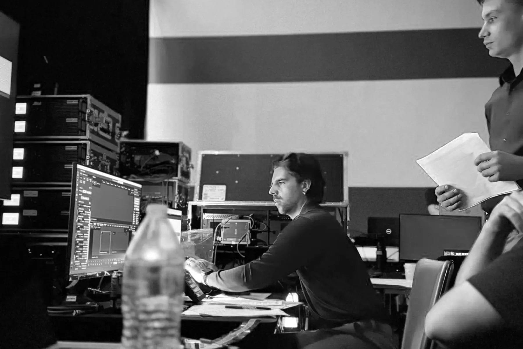 Two people in a control room work at computer screens with audio or video editing software. One is seated and focused on the monitor, while the other stands holding papers. Equipment and cables are visible in the background.