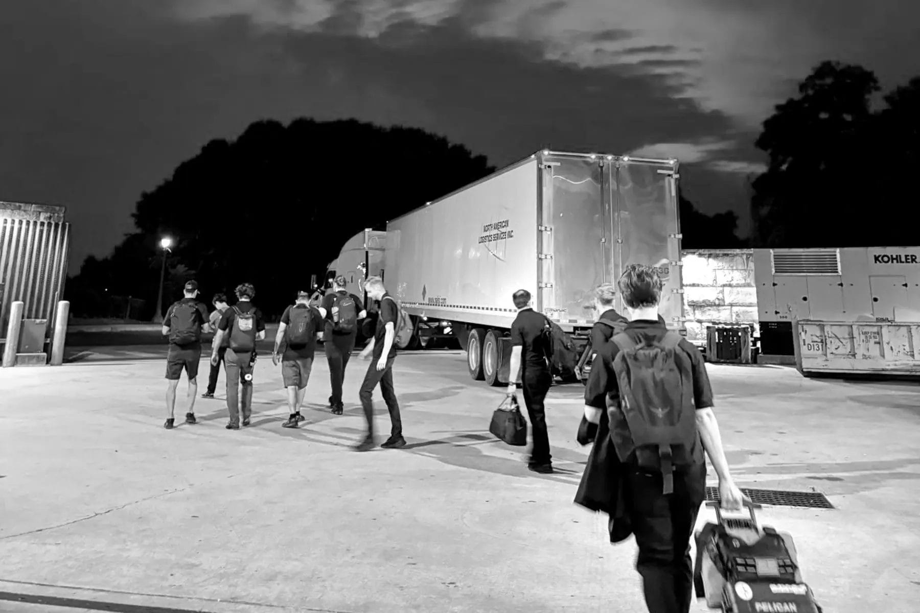 A group of people with backpacks and bags walk across a parking lot at dusk near large trucks and trees, under a cloudy sky.